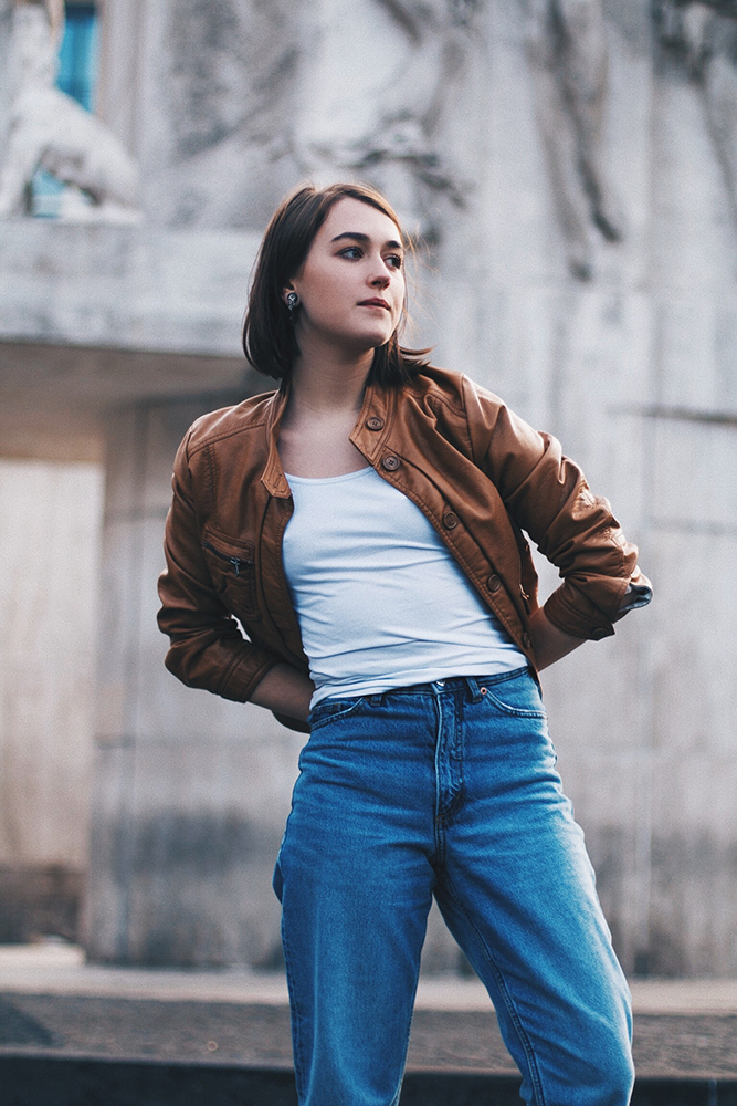 Woman standing in jeans, white tee-shirt and brown jacket.